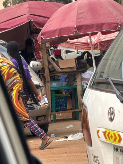 The image depicts a street scene in Orji, Nigeria, likely a marketplace or roadside stall area. Two large, red umbrellas provide shade over a wooden structure. Underneath the umbrellas, a stall is set up with various items, including packaged goods and boxes. Several individuals are visible, some obscured by the umbrellas and vehicles. One person in the foreground, wearing a purple shirt and plaid pants, walks with a plastic bag in their hand. Another person, seen from behind, has long braided hair. A white van is parked in the foreground, with its rear visible, displaying reflective safety markings. The ground is covered in reddish-brown dirt, with some scattered plastic debris. The overall impression is a busy, informal commercial setting.