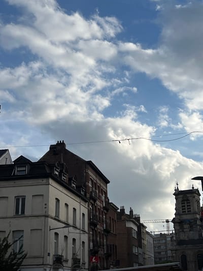 The image captures a street scene in Saint-Josse-ten-Noode - Sint-Joost-ten-Node, Belgium, under a dramatic sky filled with white and gray clouds. The perspective is looking upwards, focusing on the architecture of multi-story buildings and the sky above.
On the left, a light-colored building with a dark mansard roof dominates the foreground. It features multiple windows with dark frames on its facade, some of which are clearly visible. Adjacent to it, to the right, is a taller building constructed of dark red brick with numerous windows and balconies, characteristic of older European city architecture. Further to the right, a church tower with arched windows and a cross at its apex is visible, its stone facade weathered. In the middle ground, several other buildings of varying heights and architectural styles line the street, including one with a lighter facade and a darker, steeply pitched roof.
Stretched across the frame, at about mid-height between the buildings and the clouds, are dark wires, one of which has a red string with a small object attached, possibly part of a temporary decoration or lighting. A large construction crane is visible in the background, suggesting ongoing development or renovation in the area. The overall impression is of a dense urban environment with a mix of historical and more modern structures, bathed in the diffuse light of a partly cloudy day. There are no people visible in the image.