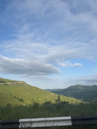 The image captures a scenic view of a mountainous landscape in Aghvani, Armenia. The scene is dominated by rolling green hills that stretch into the distance, covered in lush grass. Rocky outcrops and patches of trees are visible on the slopes. In the foreground, a section of a roadside barrier with black and white markings runs along the right side of the frame, suggesting a road carved into the mountainside.
The sky above is a vibrant blue, dotted with a mix of soft, wispy white clouds and larger, denser gray clouds. The lighting suggests it is daytime, possibly late morning or early afternoon, with diffused sunlight filtering through the clouds. The overall atmosphere is serene and natural, evoking a sense of peace and the grandeur of the natural world. There is no visible text in the image. The composition is a landscape with a focus on the natural beauty of the mountains and the sky.