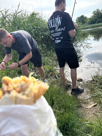 The image displays an outdoor scene featuring two men by a body of water, located in Zamfir, Bulgaria. In the immediate, blurred foreground, a food item, possibly a wrap or sandwich with visible fillings like meat and cheese, is present, partially enclosed in a white wrapper.

On the left side of the frame, a man with short, light brown hair, wearing a grey t-shirt, black shorts, a black wristband on his left arm, and a dark digital watch on his left wrist, is bent over. He is focused on manipulating thin strands of green vegetation with both hands, appearing to prepare something from the grass near the ground. A small white earbud or hearing aid is visible in his left ear.

To the right and slightly behind him, a second man stands with his back to the viewer, holding a red fishing rod extended over the water. He wears a black t-shirt with extensive white text on the back, which reads: "1987 ESTD", "FOLLOW our Heart", followed by smaller text: "TRUST IN YOUR PASSION AND INSTINCTS, FOR THEY LEAD YOU TO YOUR TRUE PURPOSE AND HAPPINESS", and below that "FAITH HOPE JOY" above a barcode-like graphic, and "MADE IN STREETS". He is also dressed in black shorts and dark athletic shoes.

The environment consists of abundant tall green reeds and grass growing along the muddy bank of a calm body of water, likely a river or lake. The water's surface reflects the overcast sky and green trees that line the opposite bank. A worn, muddy path is visible under the men's feet near the water's edge. The sky above is largely cloudy, indicating a daytime setting.