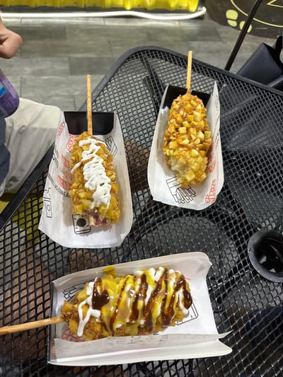 The image shows three Korean corn dogs on a black metal table. Each corn dog is on a stick and served in a white paper holder. One corn dog is coated in potato cubes and drizzled with a white sauce, another is coated in potato cubes only, and the last is coated in potato cubes and drizzled with yellow and brown sauces. The background includes a blurred glimpse of what appears to be a person’s hand and a beverage. Given the brand visible on the paper holder, the location is likely in Veracruz City, Mexico.