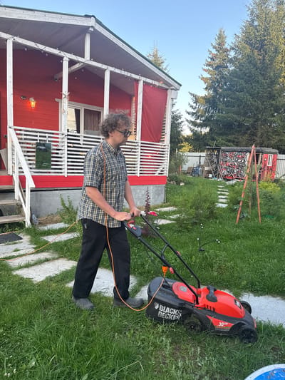 A man with curly hair and glasses, wearing a plaid shirt and black pants, is mowing the lawn in front of a red wooden house. He is pushing an orange and black electric lawnmower with a visible cord. The house has a white porch with a red railing and is decorated with string lights. The lawn is green and slightly overgrown in places. In the background, there are evergreen trees and a small shed covered in graffiti. The scene appears to be a residential yard in Lyady, Russia, on a clear day with soft lighting.