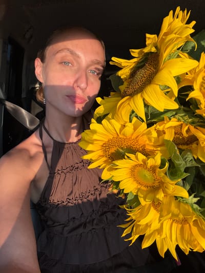 This is a close-up, top-down, portrait-style photograph taken outdoors, likely in Yanaul, Russia. The primary subjects are a young woman and a bouquet of sunflowers. The woman is lying down, her head and shoulders prominently featured in the lower portion of the frame. She has light skin, light blue eyes, and light brown hair pulled back. Her gaze is directed upwards, and her lips are slightly parted, suggesting a relaxed or contemplative mood. A shadow from the sunflowers falls diagonally across her face. She is wearing a brown, ruffled halter top.

The sunflowers dominate the upper and right portions of the image. Their large, yellow petals and brown centers are vividly captured, with some petals appearing slightly wilted. The bouquet is lush and full, with green leaves interspersed among the blooms.

The setting appears to be outdoors, with natural light indicating it is likely daytime, possibly late afternoon due to the warm, angled sunlight casting shadows. The background is dark and indistinct, suggesting the subjects are either in a shaded area or against a dark surface, possibly a car interior or a dark fabric. A gray seatbelt is visible across the woman's chest. There is no visible text in the image.