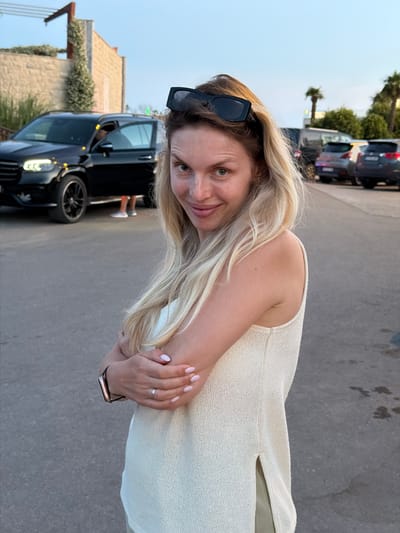 A woman with blonde hair, wearing sunglasses on her head and a cream-colored sleeveless top, stands with her arms crossed in what appears to be a parking lot in Zagora, Montenegro. Behind her, a black SUV is parked with its driver's side door open. Further in the background, other cars are visible, along with palm trees and a stone building. The sky is a pale blue, indicating it is either early morning or late evening.