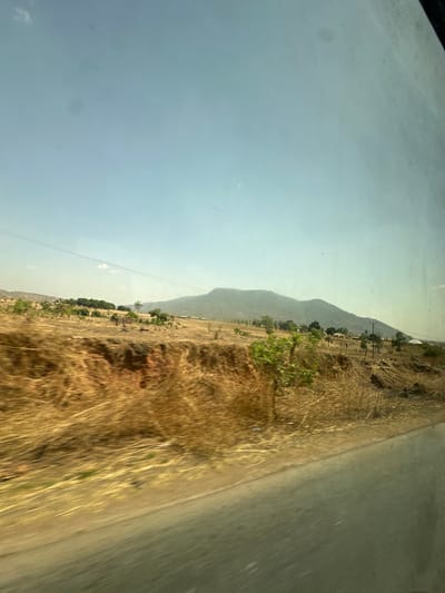 The image depicts an arid, rural landscape in the vicinity of Nandu, Nigeria, captured from the perspective of a moving vehicle. The lower right foreground shows a blurred section of a grey paved road. Adjacent to the road, an embankment features dry, golden-brown grass and scrub vegetation, with exposed reddish-brown soil visible beneath. Sparse green shrubs are present in the foreground and midground. The midground extends into broad, dry fields with scattered trees and distant, low-lying structures, indicative of human habitation. A single utility line traverses the upper midground horizontally. A large, hazy mountain range forms the horizon in the background, set against a clear, pale blue sky. No individuals or specific activities are discernible within the scene. Reflections, consistent with an image taken through a window, are visible on the sky portion.