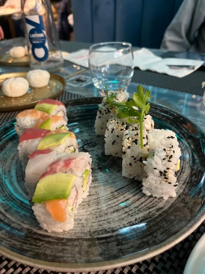 This image captures an indoor dining scene, featuring a variety of sushi rolls arranged on a dark, textured plate. The perspective is from above, looking down at the table, with the image rotated 90 degrees counter-clockwise.

The primary subjects are two distinct types of sushi rolls. In the upper portion of the plate, several pieces of uramaki (inside-out rolls) are visible, covered in white rice and generously sprinkled with black and white sesame seeds. A small sprig of fresh green herb, possibly cilantro or parsley, garnishes these rolls. In the lower part of the plate, another set of uramaki rolls is presented. These rolls feature slices of various fish, including salmon, tuna, and a white fish, along with slices of vibrant green avocado, elegantly draped over the rice. The internal fillings are not clearly visible but suggest crab stick or similar ingredients.

Adjacent to the plate, on the left side of the frame, there is a small, clear empty glass. Below it, on a small, oblong golden or brass-colored tray, rest two small, white spherical items, which appear to be miniature mozzarella balls or a similar appetizer. In the blurred background, a bottle with text is partially visible, showing "700" prominently and "S.ANNA" (or similar) below it, likely indicating a brand of water.

The setting appears to be a restaurant or eatery, suggested by the presentation of the food and the table setup, which includes a dark, woven placemat. A blue surface, possibly a wall or counter, is visible in the distant background. The lighting suggests an ambient indoor environment, suitable for a meal. No people are visible in the frame. The overall scene depicts a prepared meal of sushi and accompanying items.