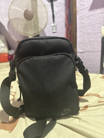 The image displays a black fanny pack, a versatile accessory designed to be worn around the waist or across the body. The fanny pack has multiple compartments, with a prominent main compartment and a smaller one on the side. It features adjustable straps with sturdy plastic buckles, indicating its functionality for securing items.

The setting appears to be indoors, possibly a bedroom or living area, given the presence of a bed with a patterned comforter and a wall with textured wallpaper. A white mesh fan is visible in the background, suggesting a warm climate, which aligns with the location context of Manila, Philippines. The lighting suggests it could be daytime, with light casting shadows. There are no people visible in the image.

The fanny pack is the primary subject, and its design suggests it is practical for everyday use, travel, or outdoor activities. The image is a close-up, focusing on the details of the fanny pack and its accessories. No visible text is present on the fanny pack or in its immediate surroundings.