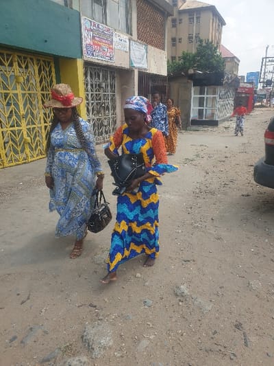 Several women stand on a dusty road. The woman in the foreground wears a light blue patterned dress, a straw hat, and carries a purse. Behind her, a woman in a blue and yellow patterned dress also carries a purse. Additional women in patterned dresses stand farther back near a building with barred windows and posters. A vehicle is partially visible at the top of the frame, with other buildings and power lines in the background.