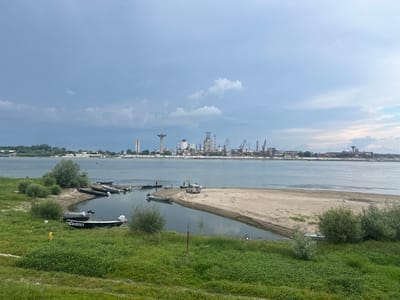 This image captures a wide view of a river scene in Nikopol, Bulgaria, featuring a lush green foreground, a sandy riverbank with inlets, the river itself, and an industrial complex on the opposite shore under a cloudy sky.
In the immediate foreground, a dense expanse of green grass and assorted low-lying shrubs covers a gently sloping embankment. To the left, a small, dark-colored motorboat with "CB 6053" stenciled in white on its bow is beached in a shallow water inlet. Next to it, a silver motorboat is also pulled onto the bank. Further into this inlet, several other dark-colored boats are visible, some partially submerged or docked.
Central to the foreground, a light-colored SUV (possibly an off-road vehicle) is parked on a broad, crescent-shaped sandy bank that extends into the water. Several individuals are present around the SUV; at least four people are discernible near the vehicle and the water's edge, one wearing a green shirt, another a blue shirt, and one a white shirt, engaged in activity near the boats or the vehicle. A dark, possibly cylindrical object or camera on a stand is positioned on the sandy bank near the SUV. The sandy area shows signs of tire tracks and foot traffic.
The midground is dominated by the wide expanse of the river, which appears calm with minimal surface disturbance, reflecting the overcast sky. The water level is visibly low, exposing additional sandy banks and shallow areas.
The background is defined by the opposite riverbank, which hosts a significant industrial complex. A series of tall, prominent structures are visible, including several water towers (one with a distinctive flared top), multiple industrial buildings with complex piping and facades, and numerous cranes (gantry cranes and dockside cranes) indicative of port operations. A tall, rectangular building is visible on the far left of the industrial skyline, and another similar structure can be seen on the far right. The sky above is largely overcast with varying shades of grey and hints of lighter blue, suggesting a cloudy day, with a few faint, distant specks, possibly birds, visible in the upper sky.