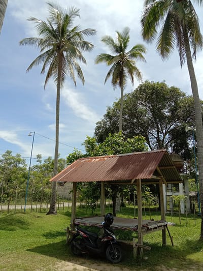 The image depicts a rural scene in Blangkarieng, Indonesia. In the foreground, a wooden gazebo with a rusted corrugated metal roof stands on a grassy area. A black motorcycle is parked next to the gazebo, with a helmet resting on its seat. Behind the gazebo, a mix of trees and vegetation is visible, including coconut palm trees. A two-story white structure with a water tank on top is partially visible behind the gazebo. A blue metal fence line and a lamppost are seen further in the background. The sky is blue with scattered clouds.