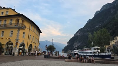 The image depicts a waterfront scene in Riva del Garda, Italy, likely in the late afternoon or early evening. On the left, a bright yellow building with the sign "HOTEL SOLE" is prominently featured. Below the hotel, an "Gelateria artigianale" with an oversized ice cream cone display is visible. Several people are seated at outdoor tables in front of the establishments.

The foreground consists of a cobblestone plaza where a group of people are sitting on a low stone wall overlooking the water. Two ferries or boats are docked in the water, one with the name "TRENTO" visible on its side. A steep, tree-covered mountain rises dramatically behind the boats and the lake. The sky is a soft blue with hints of pink and orange, suggesting the setting sun. The overall atmosphere is relaxed and picturesque, characteristic of a European lakeside town.
