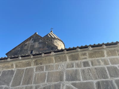A low-angle shot captures the exterior of a stone building, presumably a church or monastery. The structure features a peaked roof and a domed section topped with a cross. The stonework wall displays carved symbols, including a star-like design and crosses. The sky is clear and blue.