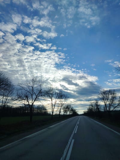 The image shows a road in Antonovo, Bulgaria. The road is gray with white lane markings. Trees, silhouetted against the sky, line either side of the road. The sky is blue with white clouds. The image depicts a landscape, specifically the road and surrounding environment. No people are visible.
