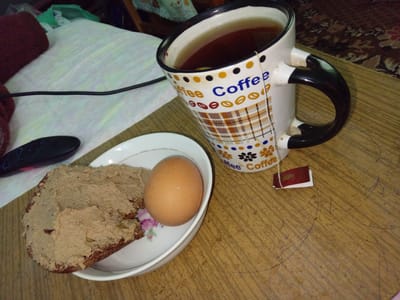 A breakfast scene unfolds on a wooden table. A white mug with a black handle, adorned with colorful polka dots and the word "Coffee" repeated, is filled with tea. A tea bag string dangles from the mug, with a small red tag attached. To the left, a white plate holds a slice of dark bread generously spread with a beige-colored pate. Adjacent to the bread is a single, light brown, hard-boiled egg. In the background, a rumpled white cloth is visible, with a dark red, knitted item to its left. A black electronic device, possibly a remote control, rests on the cloth. The wooden table shows signs of wear, with scratches and markings. The overall impression is of a simple, home-cooked breakfast. The image is set in Kitskany, Moldova.