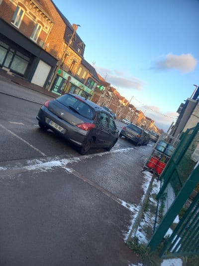 The image depicts a static street scene in Noyelles-sous-Lens, France, under a clear blue sky with scattered white clouds, suggesting daylight, possibly morning or late afternoon given the light angle. The ground, an asphalt road and adjacent sidewalk, shows visible patches of white material consistent with snow or ice.
In the foreground, a dark grey Peugeot hatchback, license plate "BD-790-NX", is parked on the street, angled slightly towards the left. Another dark-colored hatchback is parked behind it further down the street on the same side, with additional vehicles visible in the distant background.
To the left of the street, a row of multi-story buildings lines the street. The building closest to the viewer features a brick facade on its upper levels and a darker ground floor. Adjacent to it, a storefront with a vibrant green sign displays "PHARMACIE" and a green cross symbol. Further along, similar residential or commercial structures are visible.
On the right side of the image, a sidewalk partially covered in snow/ice runs parallel to the road. A dark green metal fence stands along the edge of the sidewalk. Behind this fence, a metal rack holds several gas canisters, some red and some blue/grey. No individuals or active interactions are observed within the scene.