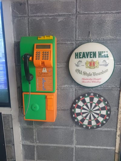This indoor shot shows a wall decorated with several items. A green and orange public telephone is mounted on the left side of the wall. To the right of the telephone is a round sign advertising "Heaven Hill Old Style Bourbon Kentucky Straight Bourbon Whiskey". Below the sign is a standard dartboard.
The telephone features a keypad, a card slot, and a handset. A small sticker with the TOT (TOT Corporation Public Company Limited) logo is affixed to the telephone.
The "Heaven Hill" sign is white with green text and trim. The dartboard is a classic design with numbered sections. The wall appears to be constructed from gray concrete blocks.
Based on the OCR reading of the TOT sticker, the location can be identified as Surat Thani City Municipality, Thailand.