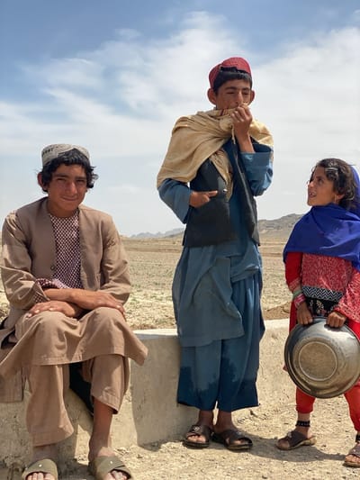 Three individuals are visible in an outdoor setting under a blue sky.
On the left, a young man sits with crossed arms, wearing a tan traditional outfit and a gray hat. He is seated on a low, concrete structure.
In the center, a taller individual stands wearing a blue outfit, a black vest, a red hat, and a light tan scarf draped over their shoulders.
To the right, a younger girl is seen holding a metal bowl. She is wearing a red dress, red pants, a blue head covering, and sandals. She appears to be looking up at the person in the center.
The backdrop features a flat, arid landscape with distant mountains visible on the horizon. The ground is dry and dusty.