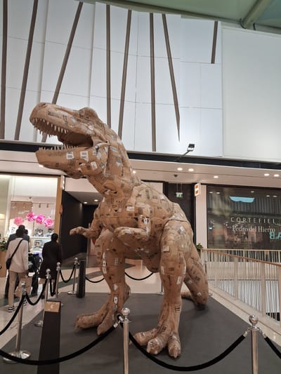 The image shows a large Tyrannosaurus Rex sculpture made of cardboard boxes. The sculpture is located inside a shopping mall, identifiable by the storefront visible in the background, "CORTEFIEL Pedro del Hierro B.A," and the architecture of the building. The sculpture is roped off with black ropes and silver stanchions.  Several people can be seen in the background near a storefront.  The wall to the left consists of white panels with brown accents. The image was taken at an angle and then rotated, distorting the view. The shopping mall could be located in the city of Amadora, Portugal, but that is impossible to confirm with the content of this image.