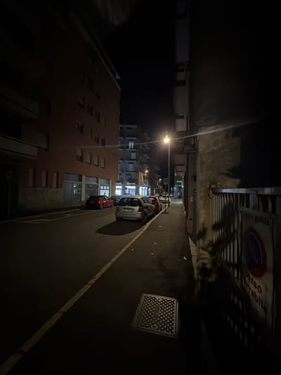 The image depicts a street scene at night in Novara, Italy. A sidewalk is visible on the right, bordered by a metal fence. A sign with text in Italian, possibly indicating a traffic regulation like "Passo Carrabile" (driveway access), is affixed to the fence. A street lamp illuminates the scene from the left side of the image, casting long shadows. Several parked cars are lined up along the street. The buildings on either side of the street are dark, with some windows showing interior lights. A sewer grate is also visible on the sidewalk in the foreground. The overall impression is one of a quiet urban street at night.