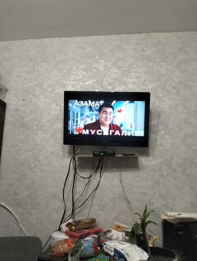 The image shows a television mounted on a textured grey wall. The TV displays a man's face with text overlays in Cyrillic. Below the television, a small wooden shelf holds electronic devices and cables. To the right of the shelf, a tangle of black cables descends the wall. In the lower portion of the image, a cluttered surface features various items including packaged food, a small potted plant, and other miscellaneous objects. The overall environment suggests an indoor residential setting.