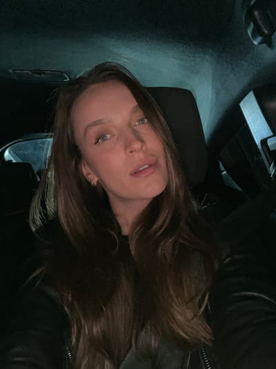 An intimate, sideways-oriented portrait features a woman with long, wavy dark brown hair. She is seen from the chest up, with her head tilted slightly back as she looks towards the camera with light-colored (likely blue or green) eyes. Her expression is calm and slightly pensive, with her lips subtly parted. Her fair skin is illuminated by a soft light source, creating highlights on her forehead, nose, and lips. She appears to be wearing a dark garment, possibly a leather jacket, visible on her right shoulder and across her chest.
The setting is dimly lit and appears to be indoors or within a vehicle, with a dark, out-of-focus background that includes faint reflections or light sources. This suggests the photo was taken at night or in a low-light environment. The confined nature of the space implies an interior location. Given the context, this scene is set in Braćeni, Montenegro. There is no visible text in the image.