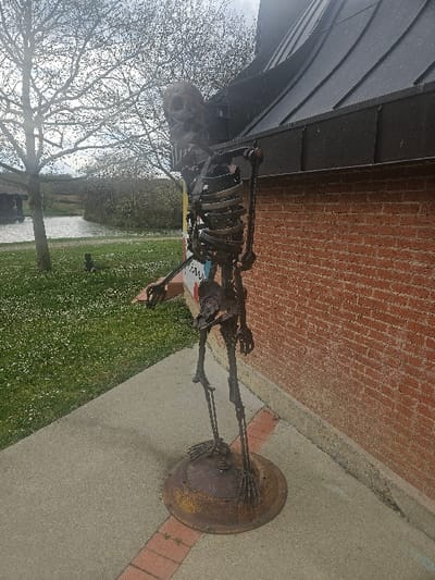 An outdoor scene in Avignonet-Lauragais, France, depicts a weathered metal skeletal sculpture positioned on a paved area. The life-sized sculpture, constructed from dark, possibly rusted, metal components, mimics a human skeleton. It stands upright on a circular, rusted metal disc base. The sculpture's right arm is slightly raised and bent at the elbow, while its left arm is bent and extends forward. The skull is tilted slightly upward and to the right.
To the sculpture's immediate right is a building with a reddish-orange brick wall, extending vertically from the ground. Above the brick wall, a dark gray sloped roof with distinct panels and a visible gutter system is present. To the sculpture's left, the paved surface, which appears to be light gray concrete, transitions into a narrow strip of reddish-brown bricks, followed by a green grassy area dotted with small white flowers.
In the midground, beyond the grassy area, a body of water, consistent with a canal or pond, is visible. The opposite bank features a prominent deciduous tree with bare branches. Further in the background, additional trees and dense green vegetation are apparent under an overcast sky.