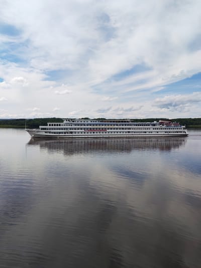 A long, white, multi-deck river cruise ship is moving on a wide, dark river. The ship has numerous windows, and red accents are visible on the upper decks. The river's surface reflects the ship and the sky, which is filled with white and gray clouds against a blue background. Trees and hills are visible on the distant bank of the river. The scene is set in Shchetinino, Russia.