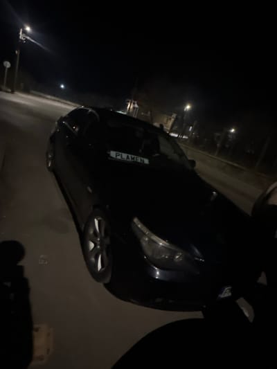 The image depicts a dark-colored sedan, likely a BMW 5 Series (E60 generation), parked on a paved surface at night in Jitnitsa, Bulgaria. The vehicle is positioned at an angle, with its front and left side visible. A white sign displaying "PLAMEN" in black capital letters is visible on the dashboard behind the windshield. Multi-spoke alloy wheels are visible on the front left side. A partial, illegible European license plate is present on the front bumper. The scene is illuminated by streetlights, with one prominent streetlight on a pole in the upper left background and other ambient lights in the distance. The environment consists of a road or parking area, with faint outlines of buildings or trees in the darker background. A dark, indistinct shadow on the lower right foreground suggests the presence of the photographer. No other individuals or specific actions are discernible.