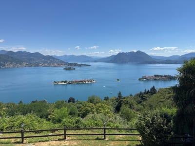 The image shows a wide, serene blue lake with several small islands dotted across its surface. The islands are covered with buildings and lush greenery, and boats can be seen navigating the water. In the distance, mountains rise majestically, their slopes covered in trees and some settlements. The sky above is a clear, bright blue with a few wispy clouds. In the foreground, a wooden fence runs horizontally across the image, with a grassy area and dense foliage behind it. The overall impression is one of a tranquil and picturesque landscape, likely a tourist destination, showcasing the natural beauty of the region. This scene does not appear to be in Campino, Italy, as Campino is a town near Rome and is not known for having a large lake with islands and mountains like this.