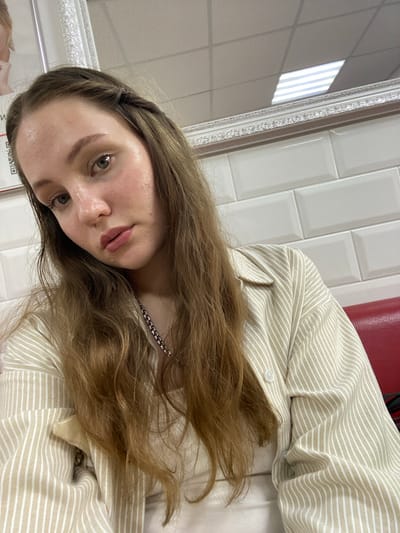 A young woman with long, light brown hair braided on one side is looking at the camera, positioned slightly to her left. She is wearing a light-colored collared shirt with thin vertical stripes and a silver chain necklace. Her skin appears slightly shiny, and she is wearing subtle makeup. Behind her, a wall is tiled with rectangular white tiles. A red surface, likely a couch or chair, is visible to her right. The ceiling is tiled with acoustic ceiling panels and features fluorescent lighting fixtures. The overall environment appears to be indoors, possibly a waiting room or a casual establishment. The exact location within Советско-Никольское, Russia, cannot be determined from the visual information alone.