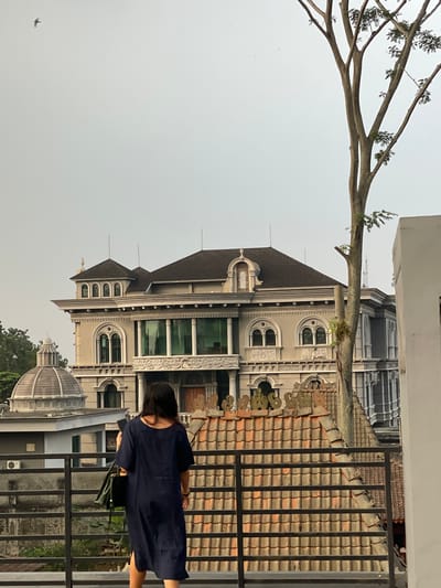 An eye-level, medium outdoor shot captures a person facing away from the camera, standing on a balcony with a dark metal railing. The individual, with shoulder-length dark hair, wears a navy blue dress and carries a dark bag over their left shoulder. In the background, a grand, ornate building with a grey facade, dark tiled roofs, and numerous windows is visible. To the left of this building, a dome-shaped structure with a grey, tiled roof sits atop another building. A tall, bare tree with some green leaves on its branches stands to the right of the main building. The sky is overcast and pale grey. The scene appears to be set in the city of Babakan Madang, Indonesia.