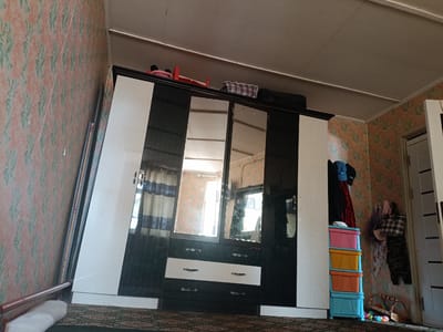 The image depicts an indoor scene, likely a bedroom, within a building in Tash-Dobo, Kyrgyzstan. The room is furnished with a large, dark wood and white wardrobe that dominates the center of the frame. The wardrobe has multiple mirrored doors, reflecting the room's interior and a window. On top of the wardrobe, a red children's toy, possibly a rocking horse, and a plaid fabric-covered object are visible.
To the left of the wardrobe, a portion of a wall with floral wallpaper in shades of pink and green is visible, along with a wooden bed frame and headboard. To the right of the wardrobe, a stack of brightly colored plastic drawers (pink, orange, yellow, and blue) is placed against the wall. Several items of clothing are hanging on hooks on the wallpapered wall next to the drawers, including a blue baseball cap, dark clothing, and bags. A white doorframe is also present on the far right. The ceiling appears to be made of white panels, with visible seams. The floor is covered with a patterned carpet in dark green and brown tones. The overall impression is of a lived-in, perhaps somewhat cluttered, domestic space.