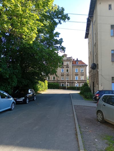The image depicts a street scene in Wałbrzych, Poland, on a sunny day. The viewpoint is from a street level, looking up a slight incline. To the left, a large, leafy green tree dominates the foreground, casting shadows on the asphalt road. Several cars are parked along the left side of the road, including a light blue car and a black car with a license plate reading "DW 4RR02".
Further up the street, on the right, is a multi-story building with a neutral-colored facade and windows. To the left of this building, and slightly further back, is another, more ornate building with a red-tiled roof and detailed architectural features, typical of older European architecture. This building appears to be an apartment block or similar residential structure.
The sky is clear and bright blue, suggesting a pleasant, possibly warm, day. The sunlight is strong, creating distinct shadows and highlights on the buildings and trees. A sidewalk runs alongside the buildings on the right. The scene appears quiet and ordinary, with no people visible. The overall impression is of a residential street in a European town.