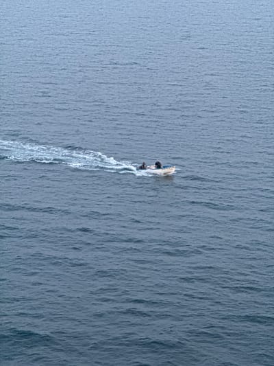 An overhead, medium shot captures a small, white motorboat with a blue trim navigating through calm, choppy, dark blue water. Two individuals are seated in the boat; one in the front and one in the rear. The boat is moving from left to right, leaving a white wake that fans out behind it. The water's surface is textured with numerous small waves, reflecting a muted, overcast sky. The scene is devoid of any landmass or structures, suggesting an open water environment. The location is the coast of Mers El Hadjadj, Algeria.