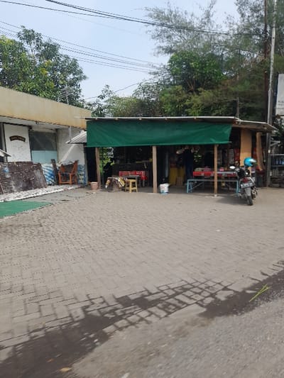 The image displays an outdoor scene in Kecamatan Gempol, Indonesia, featuring a small roadside shop under clear, sunny daylight. The shop, located on the left side of the frame, is characterized by a prominent green awning providing shade. Inside, various items are visible on shelves and racks, indicating a general store or stall. A motorbike, possibly a scooter, with a blue helmet resting on it, is parked near the shop's entrance. A few simple wooden stools or chairs are arranged in front.
To the immediate left of the shop, part of another building or an extension is visible. Overhead, utility wires crisscross against a bright, light blue sky. Lush green trees and foliage are present in the background and along the left side, suggesting a tropical or semi-rural setting.
The foreground and right side of the image show a paved road, likely constructed from cobblestones or interlocking blocks. Strong, well-defined shadows, presumably from overhead structures or trees, are cast across the road, reinforcing the impression of bright, midday or early afternoon sunlight. No specific human activity is clearly visible, presenting a static street view.
A sign above the green awning of the shop contains partially legible text, notably the Indonesian word "WARUNG," indicating a small shop or stall. Other text on this sign and on an adjacent structure is too indistinct to be read.