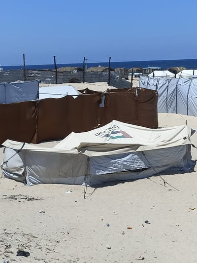 The image depicts a beach scene with several tents set up. In the foreground, a large, light-colored tent is prominent, appearing to be made of canvas or a similar material. It has a sloping roof and is supported by poles and ropes. The tent has a logo or emblem on its side, featuring a colorful design with what appears to be stripes and a shield or flag shape. Next to and behind this tent are other structures, some of which are brown canvas walls or screens, and others are white tents or shelters. In the background, more tents with thatched roofs are visible, suggesting a more permanent or semi-permanent encampment. Beyond the tents, the blue sea stretches to the horizon under a clear, bright blue sky. The ground is sandy and appears somewhat littered with debris, including small pieces of trash and some darker objects. The overall impression is that of a temporary settlement or recreational area on a beach. The lighting and clear sky suggest it is daytime, likely a sunny day. The presence of multiple tents and the sea indicates a coastal location. There is no visible activity of people in the frame, making the scene appear deserted at this moment. The visible text is on the logo of the tent in the foreground.