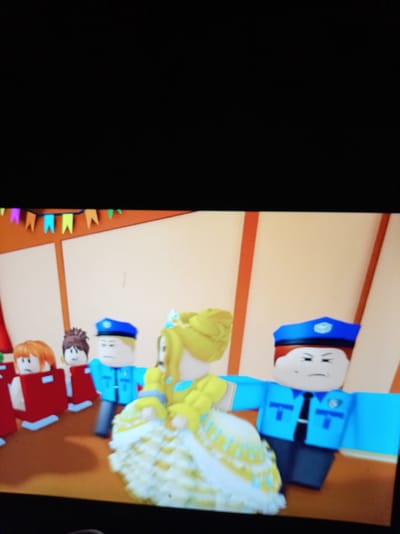 The image depicts a scene from a video game, likely Roblox, featuring stylized blocky characters. In the foreground, a character in a frilly yellow and white dress, possibly a princess or queen, is being escorted by two characters dressed as police officers in blue uniforms. The police officers have stern expressions. To the left of the princess, several other characters are visible, including one with orange hair and two in red attire. A string of colorful pennant flags is visible in the background, suggesting a festive or celebratory setting. The environment appears to be indoors, with light-colored walls and orange vertical accents. There is no indication of the city of Alatyr, Russia, in this image.