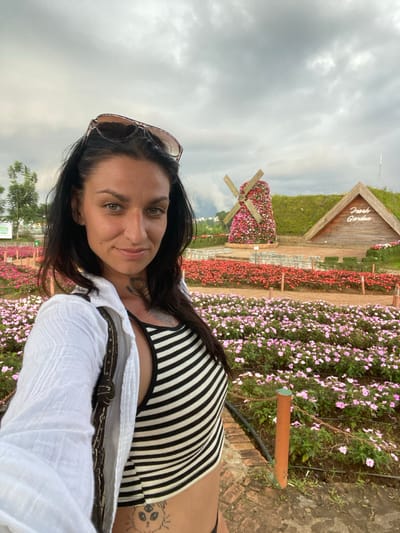 In the city of Cam Ly - Đà Lạt, Vietnam, a woman with dark hair and sunglasses perched on her head poses for a selfie outdoors. She is wearing a black and white striped crop top and an open white shirt. The background features a vibrant flower garden with rows of pink and red flowers, a decorative windmill also covered in flowers, and a wooden structure with the words "Fresh Garden" visible. The sky above is overcast.