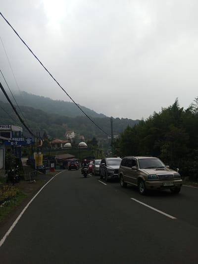 The image shows a road in Mae Chan District, Thailand, on an overcast day. A tan SUV is driving on the right side of the road, followed by a grey SUV. Other vehicles, including a motorcycle, are further ahead. A building marked "POLISI" (police) is visible on the left side of the road, alongside vegetation. A forested hillside is visible in the background, shrouded in mist. Power lines stretch across the top left of the frame.