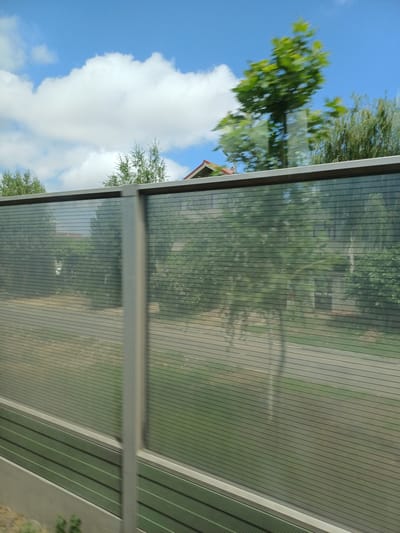 The image, captured on a sunny day in Lumina, Romania, offers a glimpse of a roadside environment characterized by a sound barrier. The barrier, constructed from transparent panels with horizontal striations, runs horizontally across the frame, partially obscuring the view behind it. A vertical silver post bisects the barrier in the foreground.

Beyond the sound barrier, the scene suggests a residential area. The blurry reflections on the transparent panels indicate motion, likely from a vehicle traveling alongside the barrier. Through the barrier, glimpses of houses with red-tiled roofs can be seen nestled amongst lush green trees. The foliage suggests a verdant, possibly suburban or semi-rural setting within Lumina. The sky above is a bright blue, dotted with fluffy white clouds, typical of fair weather. The overall impression is one of a typical urban fringe landscape, where infrastructure like sound barriers are employed to mitigate noise pollution.