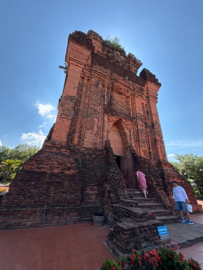 This is a low-angle, wide shot of the Thap Nhan Cham towers, a historical monument located in Tuy Hoa City, Phu Yen Province, Vietnam. The towers are constructed of red brick and are ancient Cham structures. The sky is blue with scattered white clouds.

In the foreground, on the right side of the image, a man in a white long-sleeved shirt, blue shorts, and blue athletic shoes stands on the brick steps leading up to one of the towers. He is holding a white hat in his left hand. To his left, and slightly further up the stairs, a person wearing a pink dress is ascending the steps. To the left of the stairs, a trash can is visible near the base of the tower. In the far foreground, a bed of red flowers is blooming. A blue sign with white text is also present on the stairs.

The towers themselves dominate the center and left of the image. They show signs of age with some wear and tear on the brickwork, and vegetation is growing in various crevices. The base of the tower is wider and more rugged than the upper portions. To the left of the tower, palm trees and other green foliage are visible. The ground in front of the towers is paved with reddish-brown square tiles.