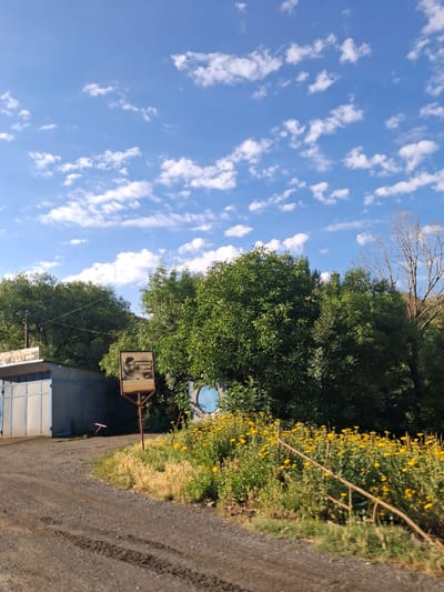 A dirt road curves past a field of yellow flowers and lush green trees under a blue sky with scattered white clouds. To the left, a corrugated metal building with blue trim and a large gate is visible. A signpost with a picture and text stands near the building and the flower field. The scene appears to be a rural or semi-rural area, possibly on the outskirts of Vayk, Armenia.