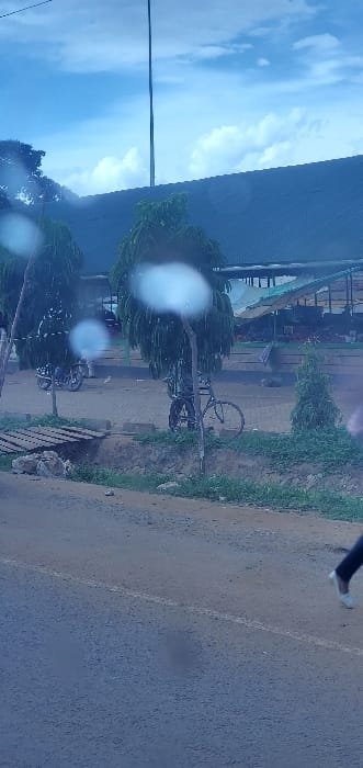 The image depicts an outdoor roadside scene in Mua ward, Kenya, viewed from a slightly elevated perspective, possibly from a vehicle, with visible water droplets or lens flares obstructing parts of the view. The foreground features an asphalt road with a faint white line on its right side. To the left of the road, a dirt shoulder with green vegetation leads to a rudimentary wooden bridge structure.
In the midground, a paved area behind the shoulder hosts several objects. Two motorcycles are parked; one on the far left has a person seated on it, and another is partially visible behind trees. A bicycle is parked upright, leaning against one of several green-leafed trees. Concrete curb stones delineate the paved area.
The background is dominated by a long, low building with a dark-colored corrugated roof. Beneath this roof, multiple market stalls with light-colored awnings or umbrellas are visible, suggesting commercial activity. A tall, slender pole, possibly an antenna or utility pole, extends vertically from behind the building towards the sky. The sky above is bright blue with scattered white clouds.
On the far right of the foreground, the lower legs and feet of a person, wearing dark trousers and light-colored shoes, are visible on the asphalt road, indicating movement or standing.