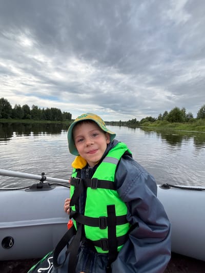 A young boy, wearing a green life vest and a patterned bucket hat, smiles at the camera while sitting in an inflatable boat. He is on a river with a treeline visible in the background. The sky is overcast. The location is Anteplevo (Okhotinskoe S/P), Russia.
