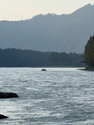 In Uznezya, Russia, a wide, choppy river flows in the foreground, with rough, white-capped waves. In the mid-ground, a small orange raft with at least three individuals aboard navigates the water. The riverbanks are lined with dense, dark green coniferous trees, forming a sharp contrast with the lighter, hazy, mountainous terrain in the background. The sky is a pale, overcast grey, with a tall, slender tower visible on the distant ridgeline, its silhouette barely discernible against the sky. Several dark rocks are visible partially submerged in the water on the left side of the frame. The overall atmosphere is muted and serene, with a sense of natural grandeur.