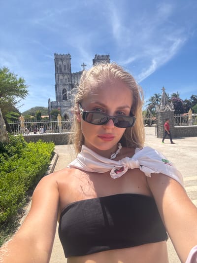 A blonde woman wearing sunglasses, a black tube top, and a white scarf tied around her shoulders takes a selfie outdoors. She is in front of a large, old stone church with two towers and a cross on top. The church appears to be made of grey stone and has Gothic architectural elements. Behind the church, there are palm trees and a clear blue sky with some wispy white clouds. To the left of the church, there is a well-maintained green hedge and a stone pathway. In the background, several people are walking around the church grounds, and one person in a red shirt is walking away from the camera near an entrance to the church. The overall setting suggests a sunny day in a historical or religious site. This location is identified as the Basilica of the Immaculate Conception in Hội Tín, Vietnam.