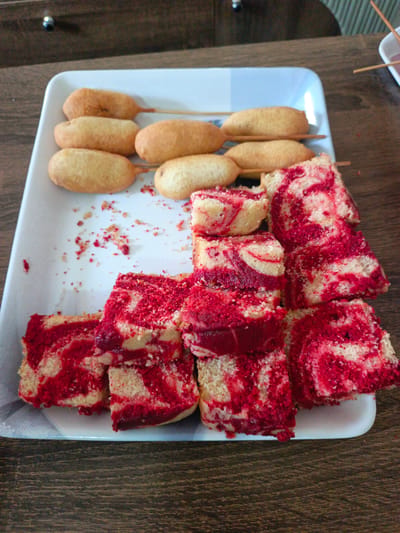 A white rectangular platter filled with food rests on a dark wooden table. The platter contains six corn dogs on skewers arranged in two rows, and a pile of sliced red velvet cake. The corn dogs are golden brown and appear to be coated in a batter. The cake is sliced into rectangular pieces, revealing a marbled pattern of red and cream-colored cake. Some red crumbs are scattered on the platter. The background is blurred, but suggests an indoor setting with wooden furniture. The image was taken in Kawo, Nigeria.