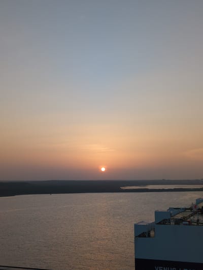 A wide shot shows a serene sunset over a calm body of water. The sky transitions from a pale blue at the top to a warm orange near the horizon, with the sun a small, bright disc positioned slightly right of center. A dark, silhouetted landmass stretches across the horizon. In the foreground, the stern of a large white cargo ship is visible, its deck structures and dark hull line with white lettering. The water reflects the soft light of the sunset, creating gentle ripples. The scene appears to be taken from a higher vantage point, possibly another vessel, overlooking the Hambantota Port in Sri Lanka. No people are visible.
