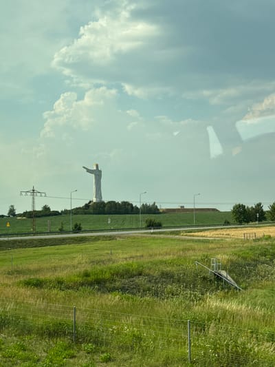 A statue of Christ the King, a massive monument in Świebodzin, Poland, stands on a hill. The statue is white with a gold crown and its arms are outstretched. The statue is surrounded by green fields and trees, with a road and power lines in the foreground. The sky is cloudy and blue. The scene is a wide shot, taken from a distance.