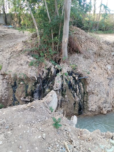 The image depicts an outdoor scene in Salar, Uzbekistan. In the foreground, a small body of water, possibly a stream or canal, flows from right to left. The banks of the water are irregular and appear to be composed of loose soil and gravel, with some rocks and debris visible. A concrete structure, possibly part of an old dam or bridge, protrudes from the bank into the water.
Behind the water and to the left, a steep, muddy embankment rises. This embankment is covered with vegetation, including trees and shrubs. Some of the vegetation appears to be growing out of the mud and rock, with roots exposed. There are also areas of dark, damp soil and what appears to be some kind of discoloration or staining on the embankment, suggesting water seepage or runoff.
Further up the embankment, a pile of loose soil and rocks is visible, likely from excavation or construction. Trees are also present in this area, with their trunks and branches extending upwards. The ground around the embankment is uneven and appears to be a mix of soil, gravel, and some vegetation.
In the background, more trees and vegetation are visible, with a wall or fence line in the far distance. The overall impression is of a natural or semi-natural environment that has been disturbed by some form of land management or construction activity. There are no people visible in the image, nor any clear evidence of recent human activity beyond the disturbed earth.